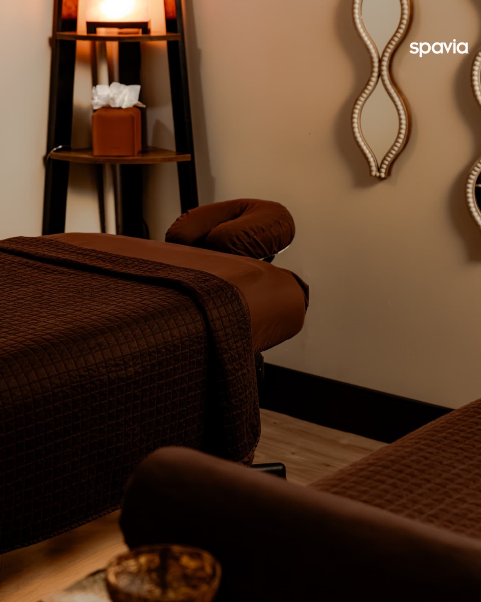 A massage table dressed in warm brown linens beside a softly glowing lamp in a Spavia treatment room